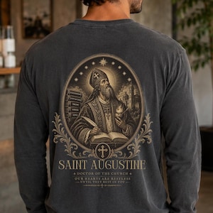 May include: A charcoal grey long-sleeve shirt featuring a detailed graphic on the back. The print depicts Saint Augustine in an oval frame with books, a quill, and a flaming heart. Text includes SAINT AUGUSTINE, DOCTOR OF THE CHURCH, and OUR HEARTS ARE RESTLESS UNTIL THEY REST IN YOU.