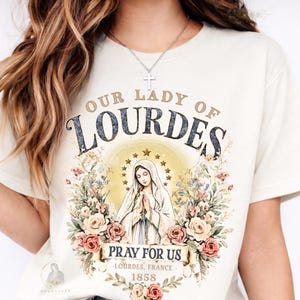 May include: Cream-colored t-shirt featuring a graphic of Our Lady of Lourdes. The design includes the text "OUR LADY OF LOURDES" above an illustration of the Virgin Mary, surrounded by flowers. The text "PRAY FOR US" and "LOURDES, FRANCE - 1858" are also present.