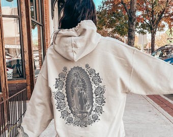 May include: A cream-colored hoodie featuring a gray illustration of a religious figure surrounded by roses. The hoodie has a drawstring hood and a kangaroo pocket. The image is centered on the back of the hoodie. The word "Guadalupe" is printed on the bottom left.