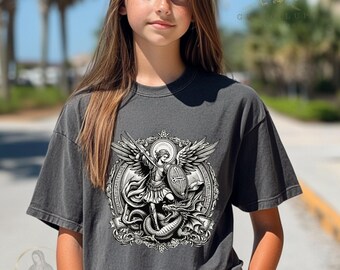 May include: Dark gray t-shirt featuring a detailed black and white graphic of an angel slaying a dragon. The design includes wings, a shield, and a sword, set within an ornate frame. The shirt has a vintage, worn look.