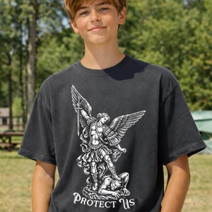 May include: Dark gray t-shirt featuring a detailed white graphic of an angel with a sword, standing over a figure. The text "PROTECT US" is below the graphic. The shirt is a crew neck style.
