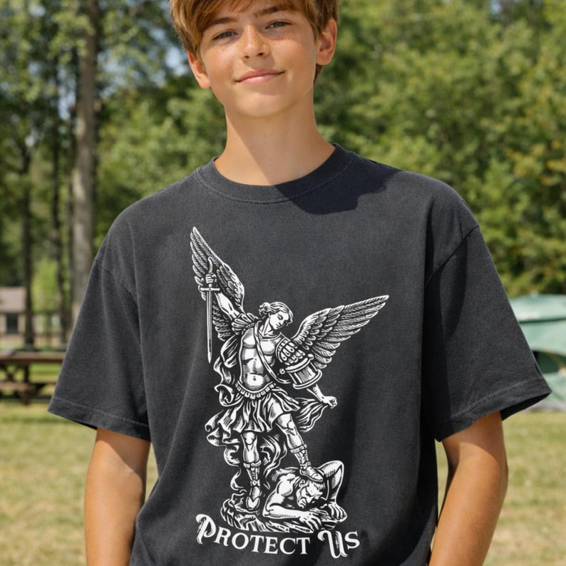 May include: Dark gray t-shirt featuring a detailed white graphic of an angel with a sword, standing over a figure. The text "PROTECT US" is below the graphic. The shirt is a crew neck style.