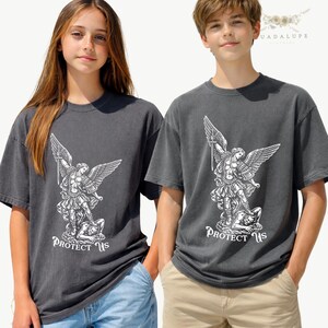 May include: Two charcoal gray t-shirts featuring a white graphic of an angel with a sword, wings, and the text "PROTECT US". The shirts are worn by two people. The shirts are made of a soft material.