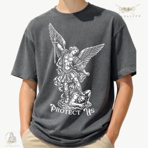 May include: Dark gray t-shirt featuring a white graphic of an angel with outstretched wings, holding a sword, and standing over a figure. The text "PROTECT US" is below the graphic. The shirt is a casual style.
