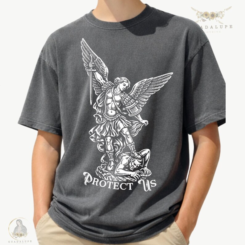 May include: Dark gray t-shirt featuring a white graphic of an angel with outstretched wings, holding a sword, and standing over a figure. The text "PROTECT US" is below the graphic. The shirt is a casual style.