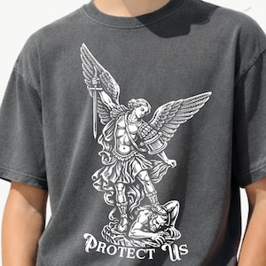 May include: Dark gray t-shirt featuring a detailed white graphic of an angel with a sword, wings, and armor, standing over a figure. The text "PROTECT US" is printed below the graphic.