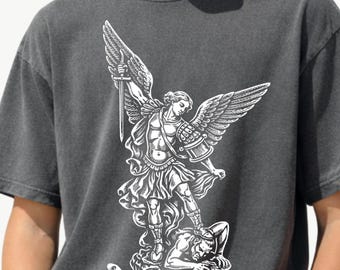 May include: Dark gray t-shirt featuring a detailed white graphic of an angel with a sword, wings, and armor, standing over a figure. The text "PROTECT US" is printed below the graphic.