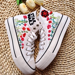May include: Pair of white high-top sneakers with black soles and white laces. The sneakers feature embroidered red flowers and green leaves. The text "STAR WARS" is embroidered on one shoe. A black and red design is on the other shoe.