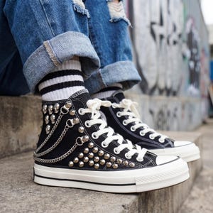 May include: Black high-top sneakers with white laces and a white rubber sole. The sneakers are adorned with silver studs, chains, and skull accents. The shoes are worn with blue jeans and striped socks.