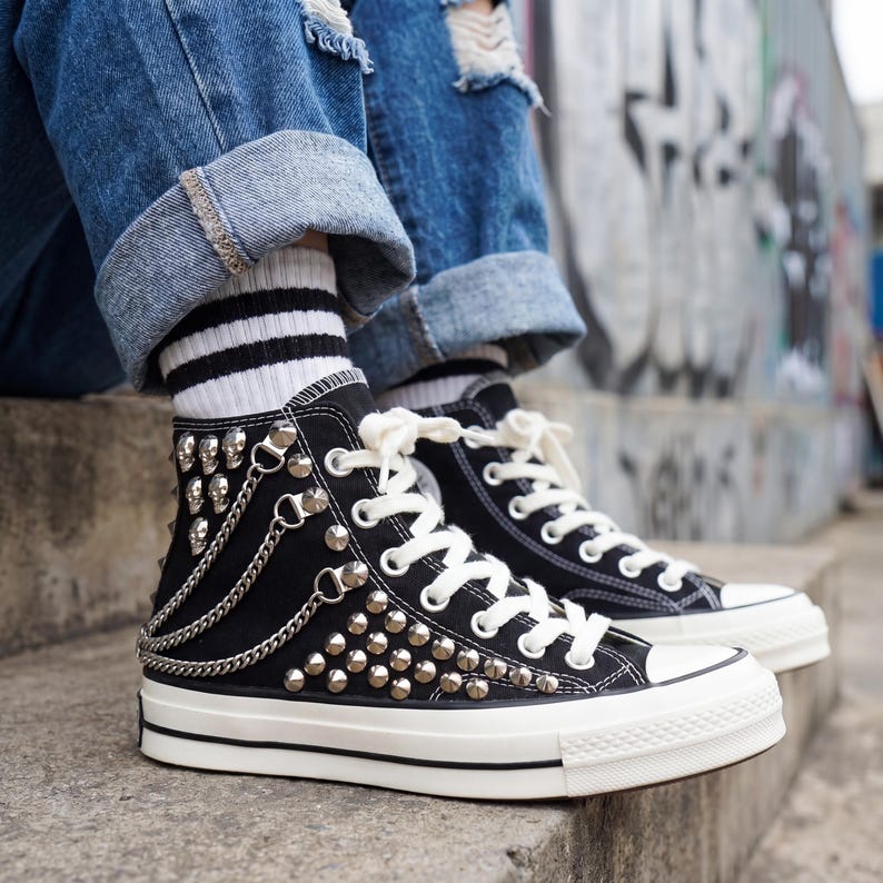 May include: Black high-top sneakers with white laces and a white rubber sole. The sneakers are adorned with silver studs, chains, and skull accents. The shoes are worn with blue jeans and striped socks.