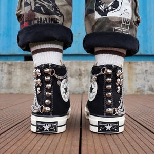 May include: Black Converse All Star high-top sneakers with silver-tone metal embellishments, including studs, chains, and charms. The sneakers have white soles and the Converse All Star logo. The shoes are worn with camouflage pants and striped socks.
