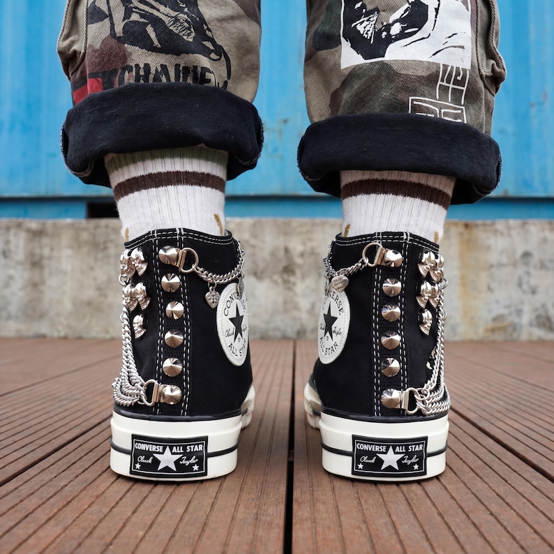 May include: Black Converse All Star high-top sneakers with silver-tone metal embellishments, including studs, chains, and charms. The sneakers have white soles and the Converse All Star logo. The shoes are worn with camouflage pants and striped socks.