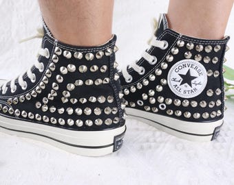 May include: A pair of black high-top canvas sneakers featuring silver-toned metal cone studs covering the outer sides. The shoes have white laces, white rubber toe caps, and a white circular Converse All Star patch on the inner ankle area.