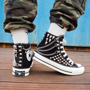 May include: Black high-top sneakers with silver studs, chains, and skull accents. The sneakers have white soles and laces, and feature the Converse All Star logo. The shoes are on a wooden surface.