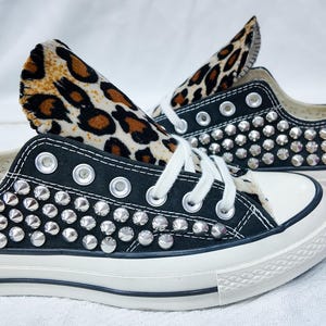 May include: Black canvas sneakers with silver stud embellishments. The shoes have white laces, a white toe cap, and a white sole. The inner lining features a leopard print design.