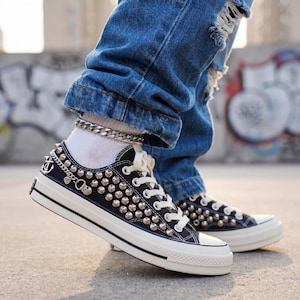 Puede incluir: Zapatillas de lona negras con tachuelas plateadas y suela de goma blanca. Las zapatillas tienen cordones blancos y un detalle de cadena plateada. Los zapatos se usan con calcetines blancos y vaqueros azules.