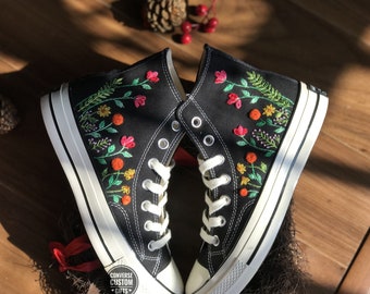 May include: Pair of black high-top sneakers with white soles and laces. The sneakers are adorned with colorful embroidered floral designs, including red, orange, and yellow flowers with green leaves. The brand name "CONVERSE CUSTOM GIFTS" is visible on the side.