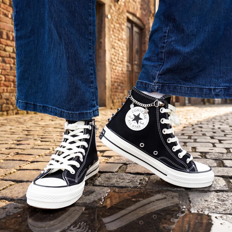 May include: Black high-top sneakers with white laces and soles. The sneakers feature the Converse All Star logo on the side and a chain with charms. The shoes are worn with blue denim jeans.