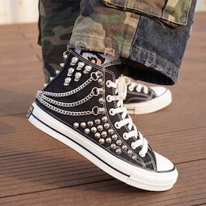 May include: Black high-top sneakers with silver studs, skull accents, and chain detailing. The sneakers have white laces, a white rubber sole, and a black canvas upper. The shoes are a bold fashion statement.