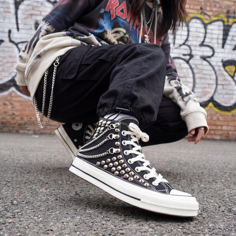 May include: Black high-top sneakers with silver studs and chain accents. The sneakers have white laces and soles. The shoes are worn with black pants.