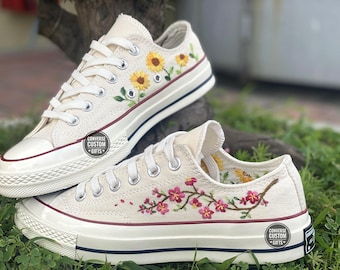 May include: A pair of white canvas Converse sneakers with custom embroidery. One shoe features a sunflower design, while the other has a cherry blossom design. Both shoes have a red stripe around the edge of the sole and a black Converse logo on the back.