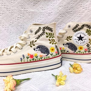 May include: Cream-colored high-top sneakers with embroidered floral and possum designs. The sneakers feature a red stripe on the midsole and the Converse All Star logo on the ankle. The embroidery includes green leaves, yellow sunflowers, and colorful flowers.