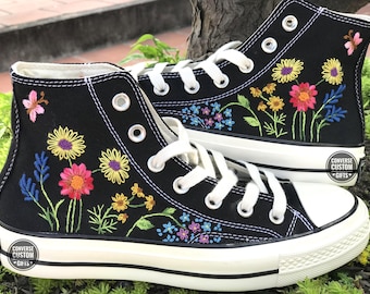 May include: Black high-top canvas sneakers with colorful embroidered flowers and leaves. The sneakers have white laces and a white rubber sole. The text "CONVERSE CUSTOM GIFTS" is printed on the side of the sneakers.