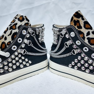 May include: Pair of black high-top sneakers with white laces and soles. The tongue features a leopard print design. The sneakers are embellished with silver studs and chain details, creating a bold, edgy aesthetic.