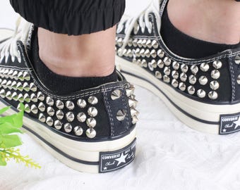 May include: Black low-top canvas sneakers featuring rows of silver-toned metal cone studs across the sides and heel. The shoes have white rubber soles with a black stripe and a Converse All Star Chuck Taylor logo patch on the back of the heel.