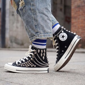 May include: Black high-top sneakers with silver studs, spikes, and chains. The sneakers have white laces, a white sole, and the Converse All Star logo on the side. The shoes are worn with blue jeans and striped socks.