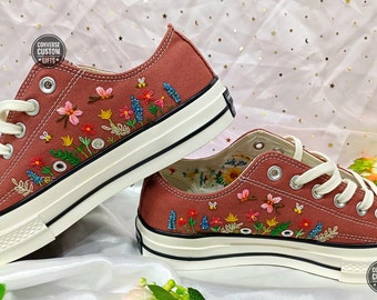 May include: A pair of brown canvas low-top sneakers with white soles and laces. The sneakers are embroidered with colorful flowers and leaves. The text "CONVERSE CUSTOM GIFTS" is on the side of each shoe.