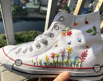 May include: White high-top Converse sneakers with colorful embroidered flowers and butterflies. The shoes have a red stripe around the sole and the text "CONVERSE CUSTOM GIFTS" on the side.