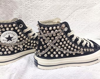 May include: Black high-top sneakers with silver metal studs and chain accents. The sneakers feature white laces, a white rubber sole, and the Converse All Star logo on the side. A stylish and edgy footwear choice.