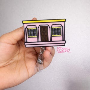 Puede incluir: Un portatarjetas rosa con un dise&ntilde;o de casa de dibujos animados. Tiene un techo amarillo, una puerta marr&oacute;n y dos ventanas con persianas negras. El portatarjetas tiene un clip de pl&aacute;stico transparente y un cord&oacute;n retr&aacute;cttil. El texto "Mary" es visible.