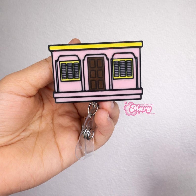 Puede incluir: Un portatarjetas rosa con un dise&ntilde;o de casa de dibujos animados. Tiene un techo amarillo, una puerta marr&oacute;n y dos ventanas con persianas negras. El portatarjetas tiene un clip de pl&aacute;stico transparente y un cord&oacute;n retr&aacute;cttil. El texto "Mary" es visible.