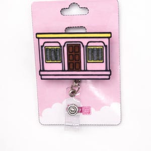 Puede incluir: Un portatarjetas retr&aacute;ctil rosa con un dise&ntilde;o de edificio rosa, puerta marr&oacute;n y ribete amarillo. El portatarjetas tiene una correa de pl&aacute;stico transparente y un clip de metal, sujeto a una tarjeta de respaldo rosa.