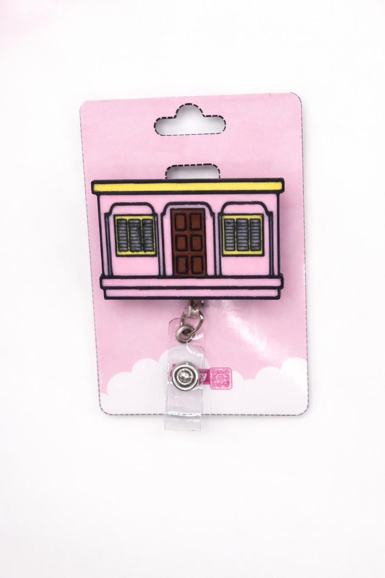 Puede incluir: Un portatarjetas retr&aacute;ctil rosa con un dise&ntilde;o de edificio rosa, puerta marr&oacute;n y ribete amarillo. El portatarjetas tiene una correa de pl&aacute;stico transparente y un clip de metal, sujeto a una tarjeta de respaldo rosa.