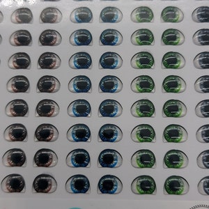 May include: A sheet of 55 pairs of doll eyes in various colours, including brown, blue, and green. The eyes are arranged in rows on a white background. The sheet is labelled "Humanizado 02 55 Pares AxL 5x6,5mm".