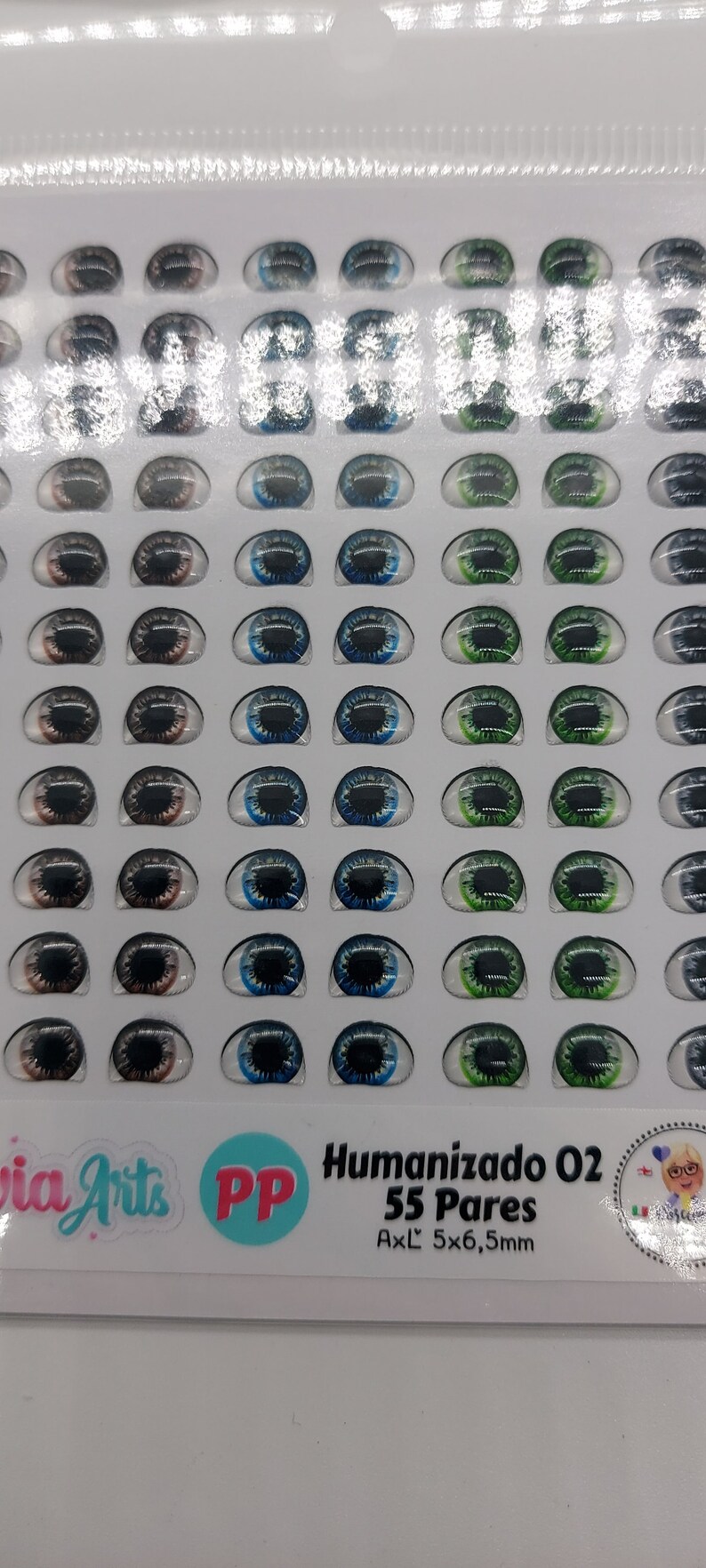 May include: A sheet of 55 pairs of doll eyes in various colours, including brown, blue, and green. The eyes are arranged in rows on a white background. The sheet is labelled "Humanizado 02 55 Pares AxL 5x6,5mm".