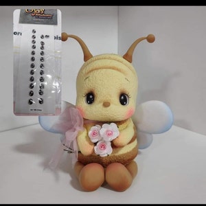 May include: A yellow bee figurine with brown stripes, antennae, and light blue wings, holding a small bouquet of white flowers with pink centers. A sheet of black oval adhesive eyes labeled Art 195 Chico is displayed in the upper left corner.