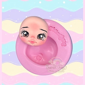 Könnte beinhalten: Eine runde, rosafarbene Silikonform mit einer vertieften kreisförmigen Aussparung und einem modellierten Puppengesicht mit großen braunen Augen, langen Wimpern und rosa Lippen. Die Form ist mit dem Text @biscuitjessy und einem Regenbogenlogo versehen.