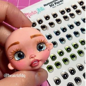 May include: A doll head with painted brown eyebrows, blue eyes, and pink cheeks. The doll head is next to a sheet of 50 pairs of doll eyes in various colours and patterns. The sheet is labelled "Humanizado 02 50 Pares Ax.C 6x8mm".