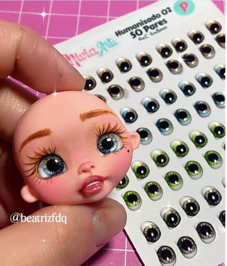 May include: A doll head with painted brown eyebrows, blue eyes, and pink cheeks. The doll head is next to a sheet of 50 pairs of doll eyes in various colours and patterns. The sheet is labelled "Humanizado 02 50 Pares Ax.C 6x8mm".