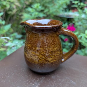 May include: A small, handmade ceramic pitcher with a dark brown glaze. The pitcher has a rounded body, a handle, and a spout for pouring. The surface has a textured appearance with lighter brown accents. The background is blurred with green foliage.