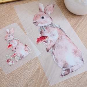 Könnte beinhalten: Zwei Bügelbilder mit Aquarell-Illustrationen von Hasen, die Pilze halten. Das größere Bild zeigt einen braunen Hasen mit einem roten Pilz, und das kleinere Bild zeigt einen braunen Hasen mit einem roten und weißen Pilz.