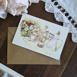Peut inclure: Illustration à l'aquarelle de souris dans un champ, avec des pissenlits et des fleurs sauvages. L'œuvre d'art est sur une carte, posée sur une enveloppe marron. Une palette d'aquarelle et des fleurs sont en arrière-plan.