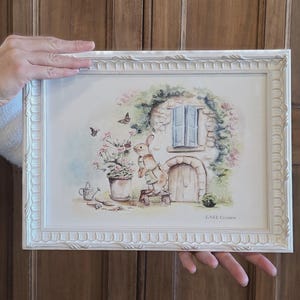 Può includere: Una stampa ad acquerello incorniciata con una scena stravagante con un coniglio, fiori e un piccolo cottage. L'opera d'arte è ambientata in una cornice bianca ornata. La stampa include il testo "E-NEE Creation".