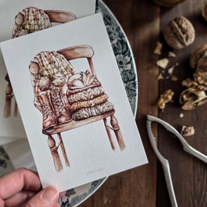 Peut inclure: Aquarelle représentant une chaise avec une couverture à carreaux, des bottes et des pulls empilés. L'œuvre d'art est sur une carte blanche, tenue dans une main, avec une assiette fleurie et des noix en arrière-plan. Thème : ambiance automnale.