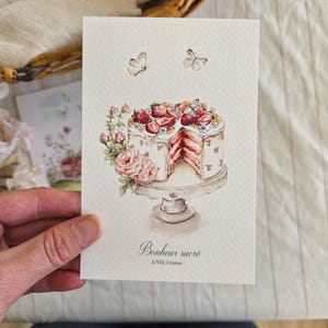 Könnte beinhalten: Aquarell-Illustration eines Kuchens auf einem Podest, verziert mit Erdbeeren, Blumen und Schmetterlingen. Der Kuchen hat ein herausgeschnittenes Stück, das das rosa Innere zeigt. Die Worte "Bonheur sucré" sind darunter geschrieben.