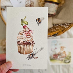 Könnte beinhalten: Aquarelldruck eines Cupcakes mit Kirsche, einer Biene und einem schlafenden Käfer. Der Druck befindet sich auf einer weißen Karte mit dem Text "E.NEE Creation". Weitere Karten und ein Glas Marmelade sind im Hintergrund zu sehen.